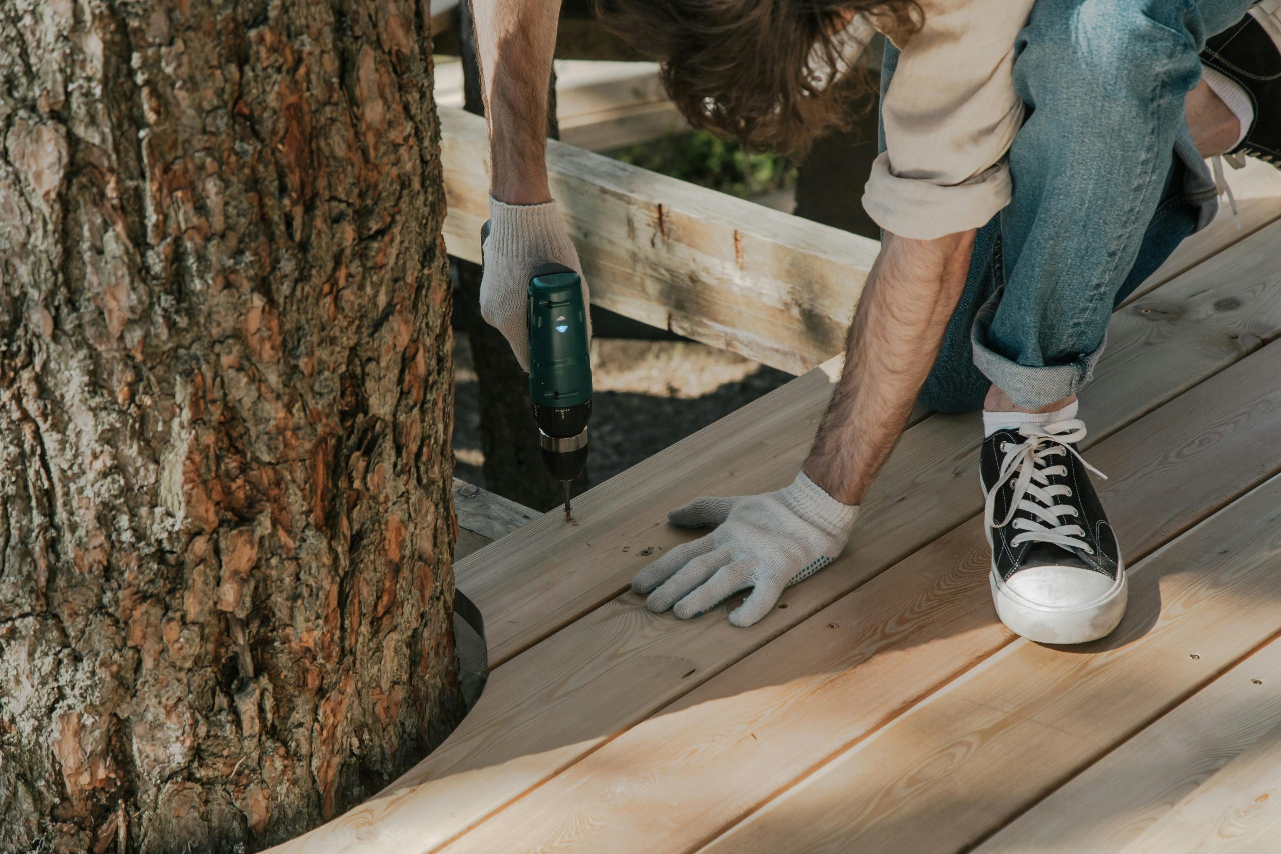 Services Worker drilling wooden planks on deck beside tree using power tool. DIY construction project.